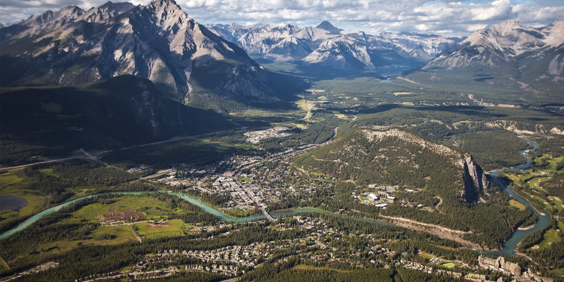 Aerial view of a mountain valley with a winding turquoise river and the town of Banff at the base surrounded by forested hills.