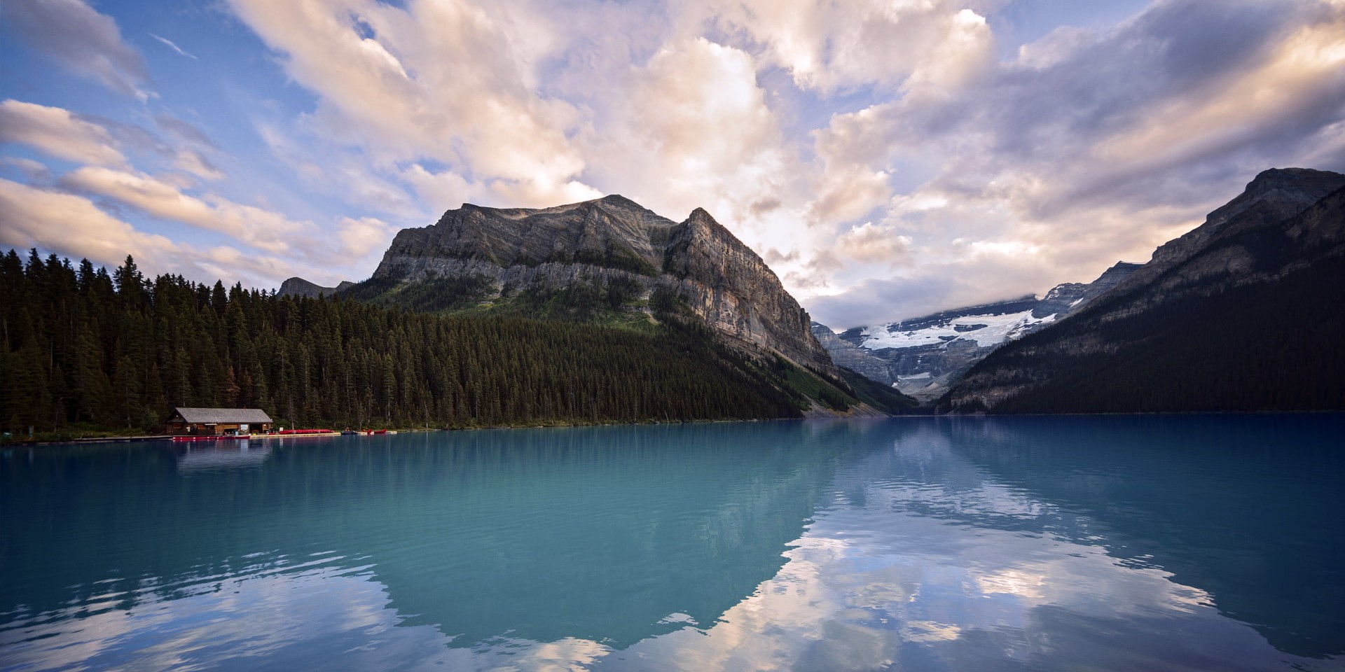 Turquoise alpine lake edged by evergreen forest and rugged mountains, with a small boathouse and red canoes along the shore under a cloudy sky.