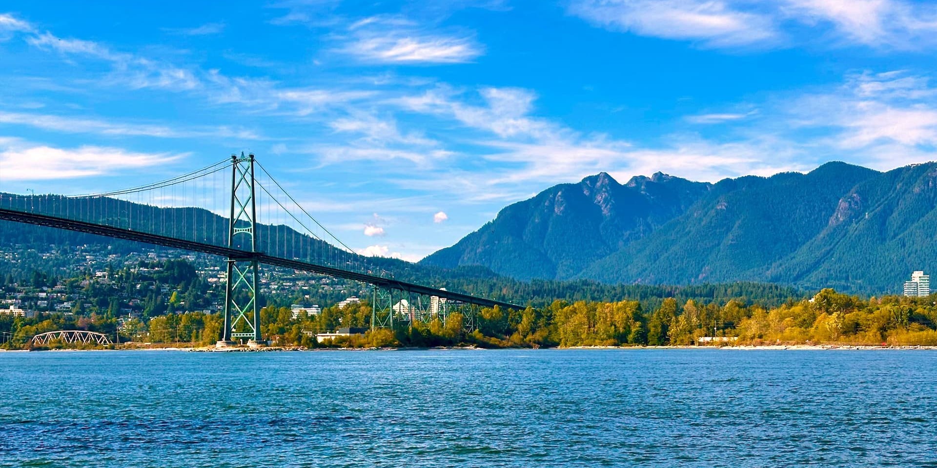 Suspension bridge spanning a blue waterway with forested hills and mountains in the background.