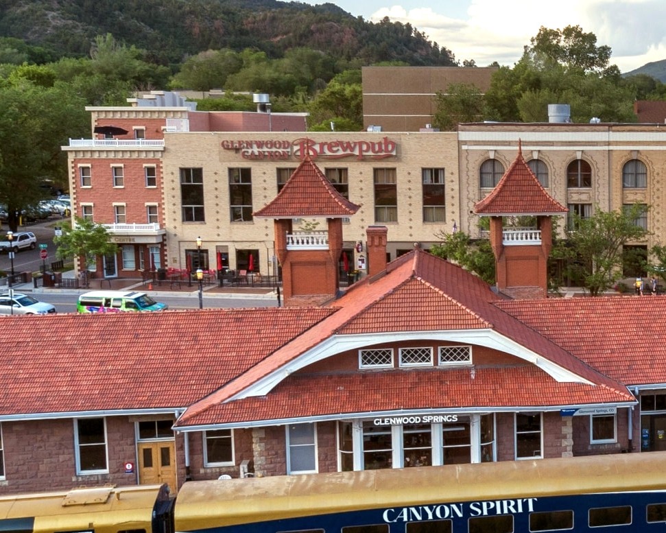 Bird's-eye view of a small town with red-tiled roofs, brick buildings, and a train labeled Canyon Spirit in the foreground. Hills frame the scene.