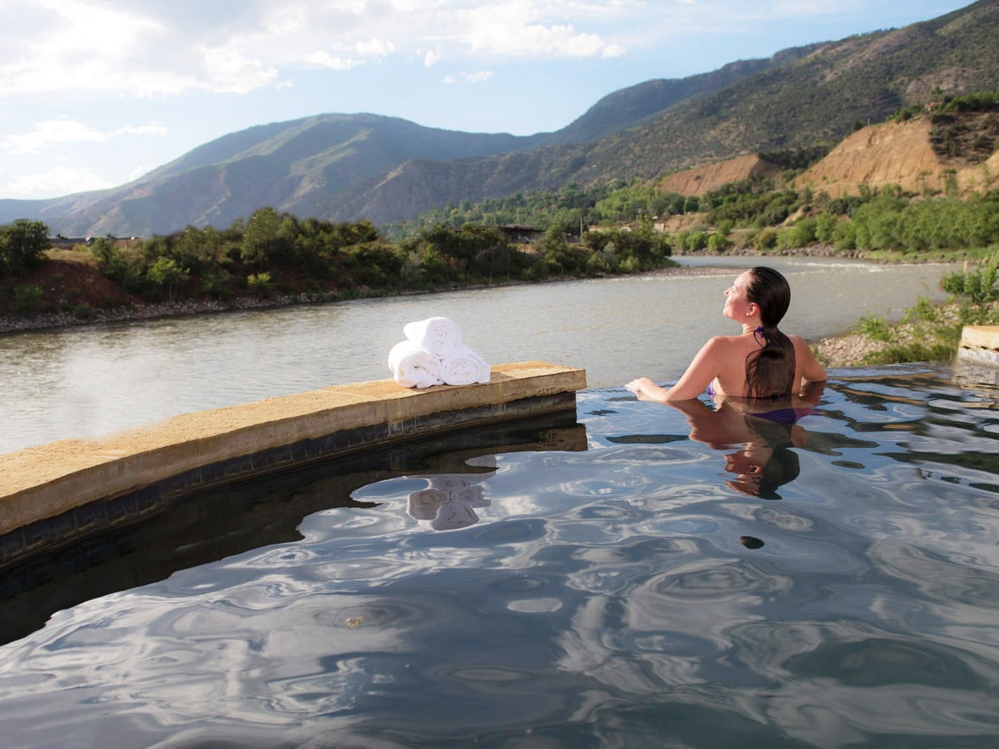 Woman reclining in an outdoor pool by a river with green mountains in the distance; white towels sit on the pool ledge.