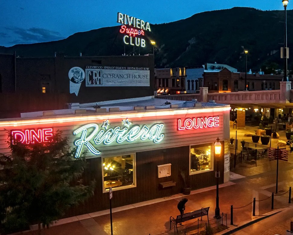 Nighttime street view of Riviera Dine & Lounge with neon signs and hillside backdrop.