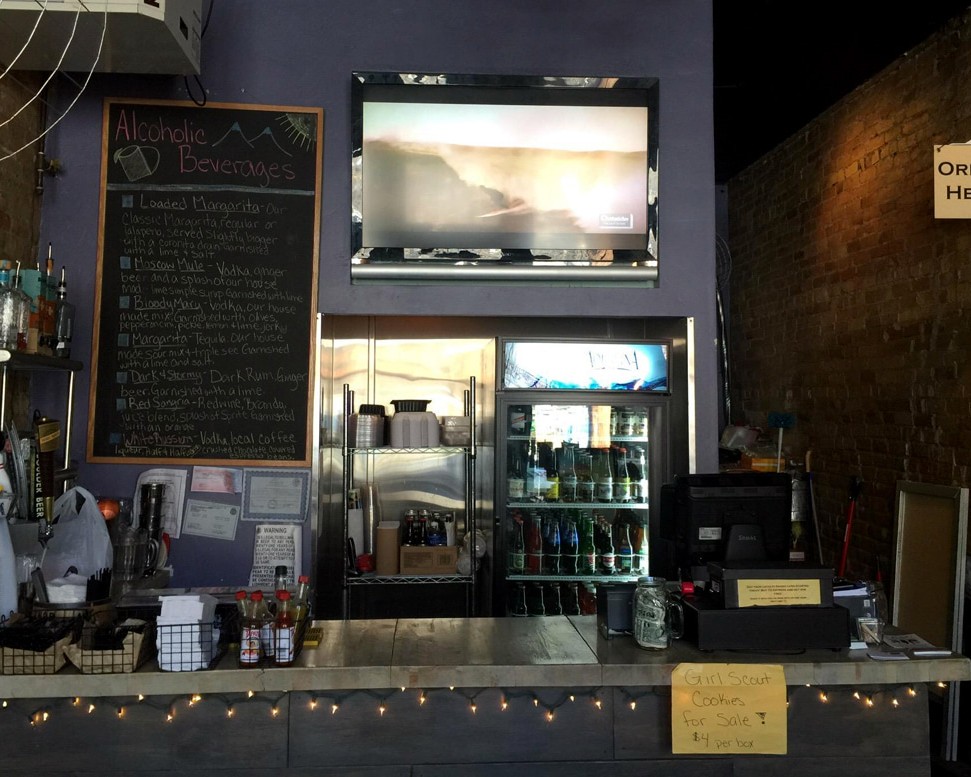 Bar counter with chalkboard menu of alcoholic beverages and a fridge behind, warm string lights along the front edge.